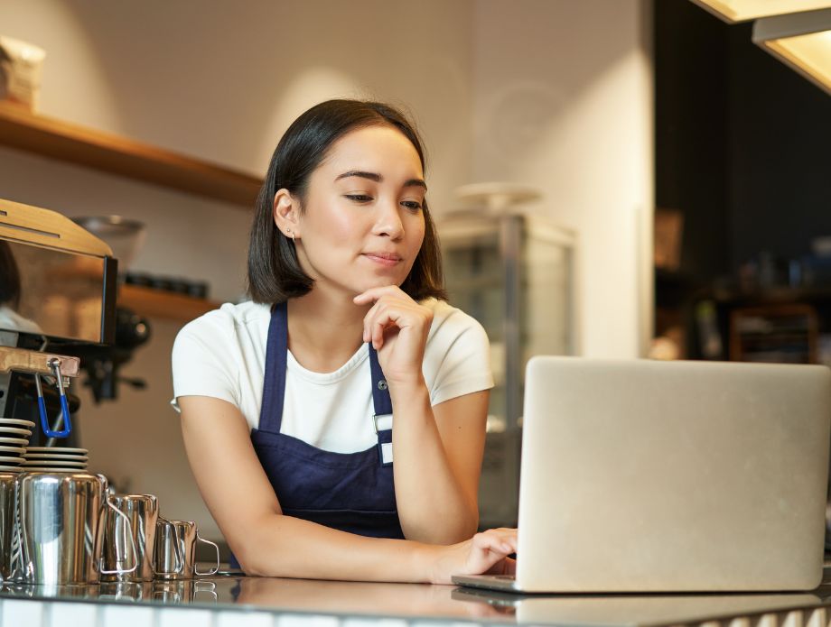 Mujer trabajando en una cafetería, usando una laptop, vestida con un delantal.